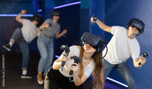 Excited modern young woman with gaming controllers in hands and VR goggles having fun with friends in virtual reality room. Concept of interactive experience.. photo