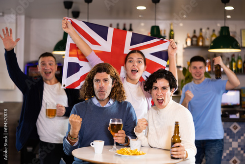Happy young adult fans waving British flag while drinking beer and watching match together in sports bar