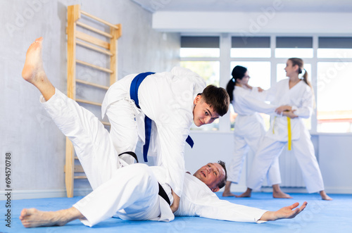 Adult man and young guy in kimonos train judo techniques in group in studio..