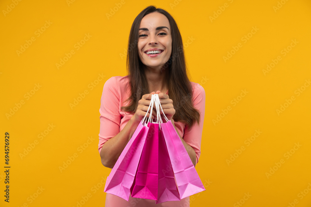 Portrait of happy smiling attractive young woman holding a bunch of bright colored pink paper shopping bags, isolated over yellow background Portrait of happy smiling attractive young woman holding a bunch of bright colored pink paper shopping bags, isolated over yellow background