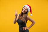 Portrait of young flirty woman in seductive black dress and Santa Claus hat posing over bright colored yellow background showing v-sign gesture