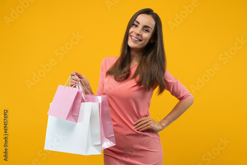 Studio portrait of attractive confident smiling young woman in stylish pastel pink dress posing over bright colored yellow background with a pile of shopping bags in hand