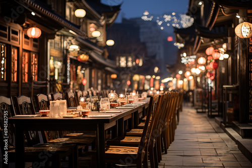 Empty outdoor dining area lined with wooden chairs and tables, illuminated by warm lantern lights in an old town street at dusk.