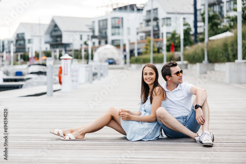 Happy man and woman sitting on a wooden pier and hugging. A young brunette woman and a stylish man in sunglasses are dressed in summer clothes. Couple in love laughing and having fun