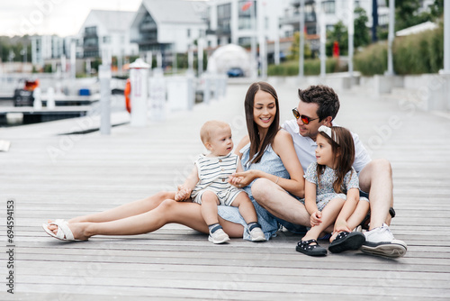 Happy family of four sitting on a wooden pier and having fun. A stylish father and a beautiful mother with their son and daughter are walking in the fresh air. Happy young family