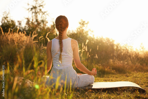 Woman practicing Padmasana on yoga mat outdoors on sunny day, back view. Lotus pose
