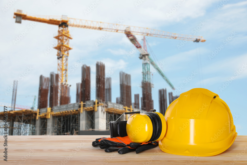 Safety equipment. Hard hat, gloves and protective headphones on wooden surface near construction site with tower crane outdoors, space for text Safety equipment. Hard hat, gloves and protective headphones on wooden surface near construction site with tower crane outdoors, space for text