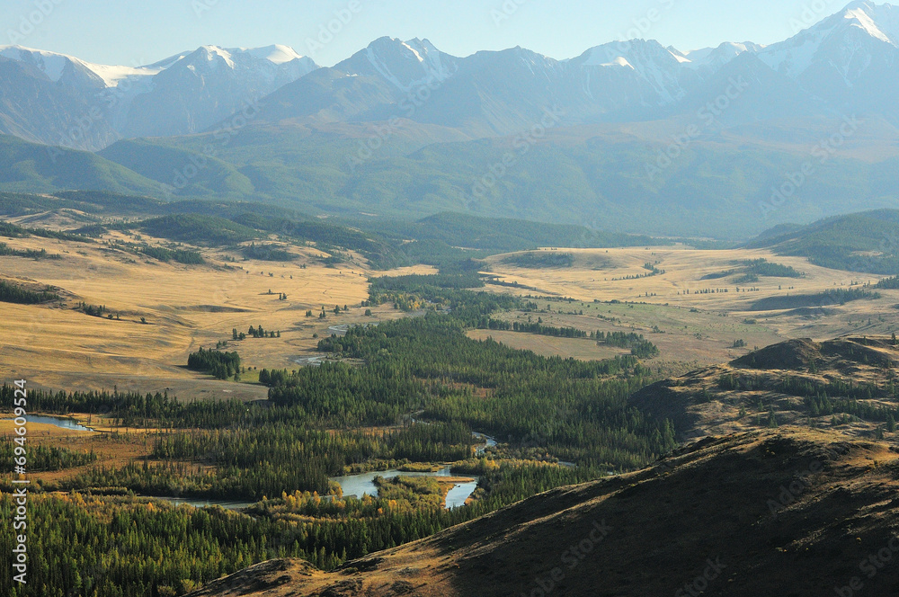 Fototapeta premium The winding bed of a beautiful river with wooded banks flows through the autumn steppe at the foot of a mountain range with snow-capped peaks in the rays of the setting sun.