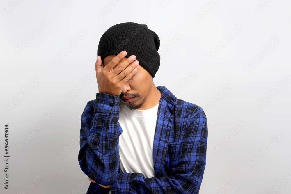 Annoyed young Asian man, dressed in beanie hat and casual shirt, is facepalming or hiding his face in frustration, feeling embarrassed due to inappropriate behavior, standing against white background Annoyed young Asian man, dressed in beanie hat and casual shirt, is facepalming or hiding his face in frustration, feeling embarrassed due to inappropriate behavior, standing against white background