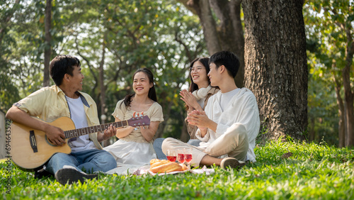 Group of cheerful Asian friends are enjoying a picnic in a park together, singing and playing guitar