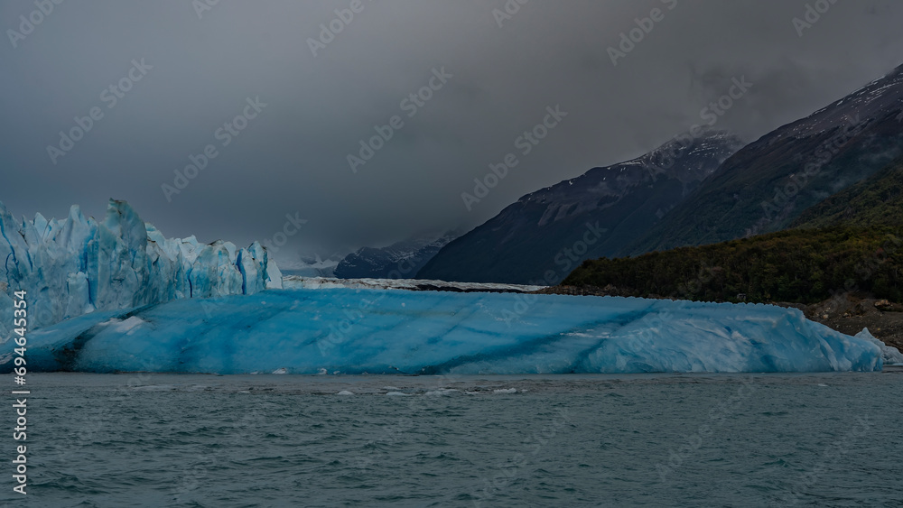 The famous Perito Moreno Glacier. A wall of cracked blue ice rises above the lake. A large breakaway iceberg floats in the water. Coastal mountains in clouds and fog. El Calafate. Argentina.