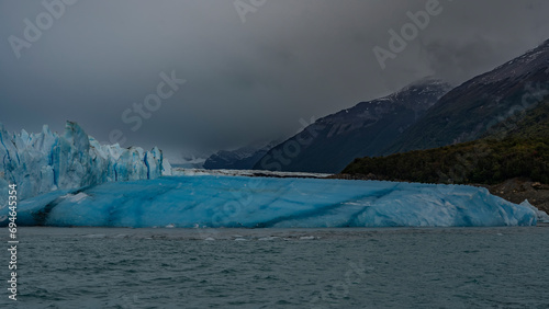 The famous Perito Moreno Glacier. A wall of cracked blue ice rises above the lake. A large breakaway iceberg floats in the water. Coastal mountains in clouds and fog. El Calafate. Argentina.