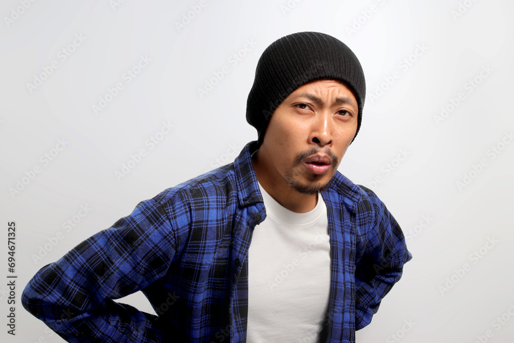 Young Asian man, dressed in a beanie hat and casual outfit, is shown holding his lower back, visibly experiencing unbearable pain due to a backache, against a white background. Young Asian man, dressed in a beanie hat and casual outfit, is shown holding his lower back, visibly experiencing unbearable pain due to a backache, against a white background.