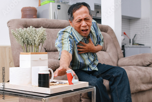 Old man suffering from asthma attack, having trouble breathing while reaching for inhaler while sitting on the sofa photo