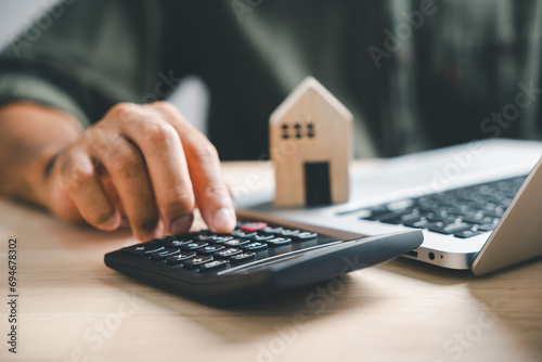 Pressing calculators hand eyes home refinance. Wooden house model, buy or rent note on desk. Saving for property purchase concept efficient mortgage payment. Tax credit analysis for financial strategy