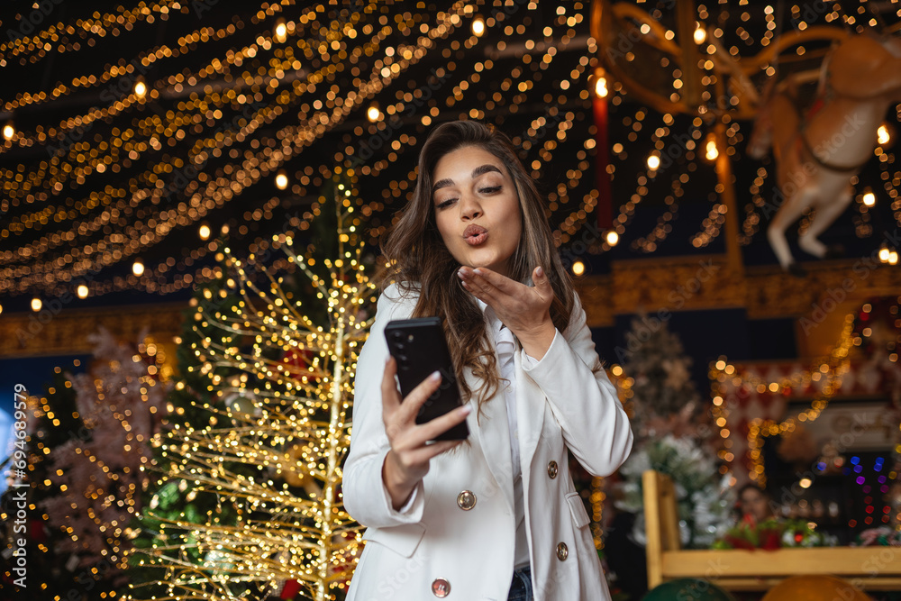 Young woman using smart phone video call talking with friend or boyfriend. Christmas tree decorated with ornament . Christmas and New Year holiday festival.