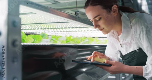 Female Biology Scientist Closely Inspecting and Analyzing Young Growing Crops. Farming Engineer Using Tablet Computer and Working in a Vertical Farm Next to Rack with Natural Eco Plants photo