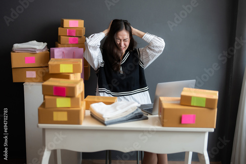 Asian female online seller, startup business owner Sad and disappointed with the sales from the website in the office. There are parcel boxes of clothes packed and ready to ship to customers.
