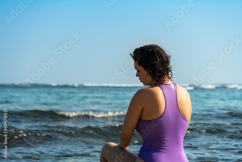 Fototapeta Naklejka Na Ścianę i Meble -  Young woman in purple swimsuit sitting if front of ocean.