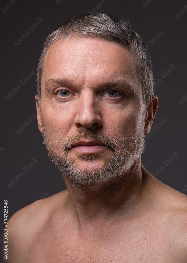 .Slightly smiling face of a middle-aged man with a beard looking at the camera. Close-up portrait of a fifty-year-old man with a naked torso against a dark background. .Slightly smiling face of a middle-aged man with a beard looking at the camera. Close-up portrait of a fifty-year-old man with a naked torso against a dark background.