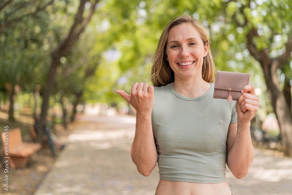 Young blonde woman holding a wallet at outdoors pointing to the side to present a product Young blonde woman holding a wallet at outdoors pointing to the side to present a product