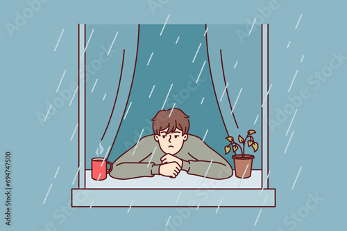 Melancholic man looks at rain from window with head on sill near coffee mug with hot drink. Melancholic guy experiences stress turning into apathy after onset of autumn due to lack of sunlight