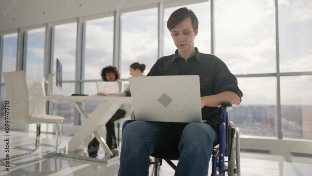 Paralyzed guy in wheelchair in office. Happy young man with physical back spine legs trauma working on laptop typing in open space workplace with colleagues. Usual life of person with special needs. Paralyzed guy in wheelchair in office. Happy young man with physical back spine legs trauma working on laptop typing in open space workplace with colleagues. Usual life of person with special needs.