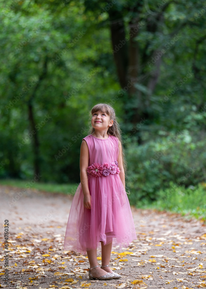 Portrait of a cute little girl 6 years old with long, dark hair. A smiling girl stands on a path with yellow leaves in the park in a beautiful pink dress and looks up with interest