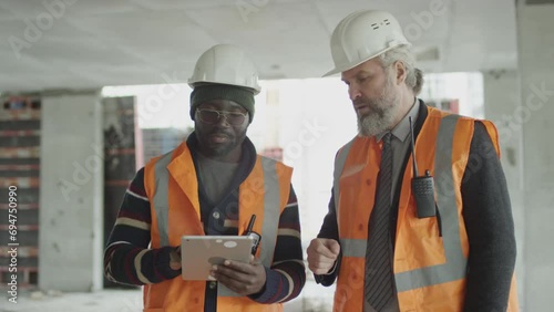 PAN medium shot of young Black male construction worker with building plan on digital tablet screen discussing issues with middle-aged Caucasian foreman both wearing safety vests and hardhats photo