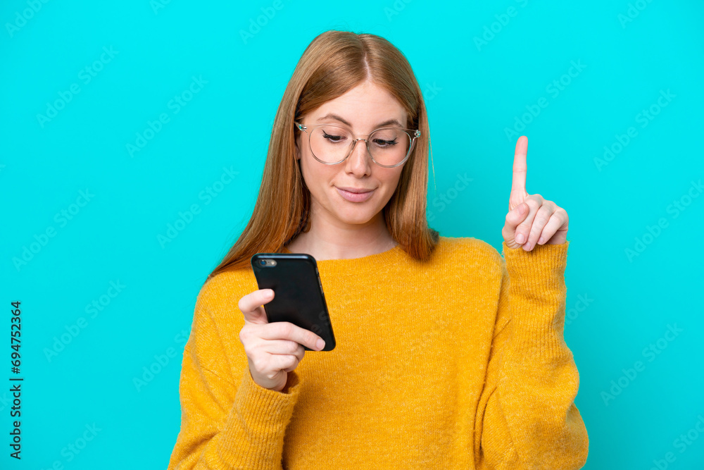 Young redhead woman isolated on blue background using mobile phone and lifting finger