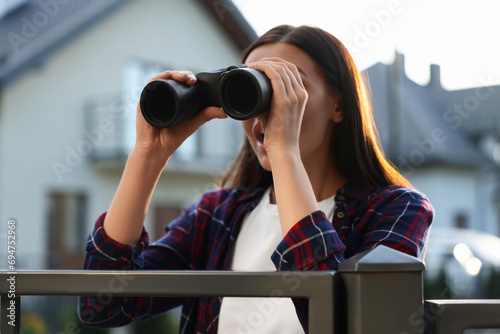 Concept of private life. Curious young woman with binoculars spying on neighbours over fence outdoors photo