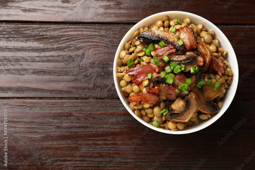 Delicious lentils with mushrooms, bacon and green onion in bowl on wooden table, top view. Space for text