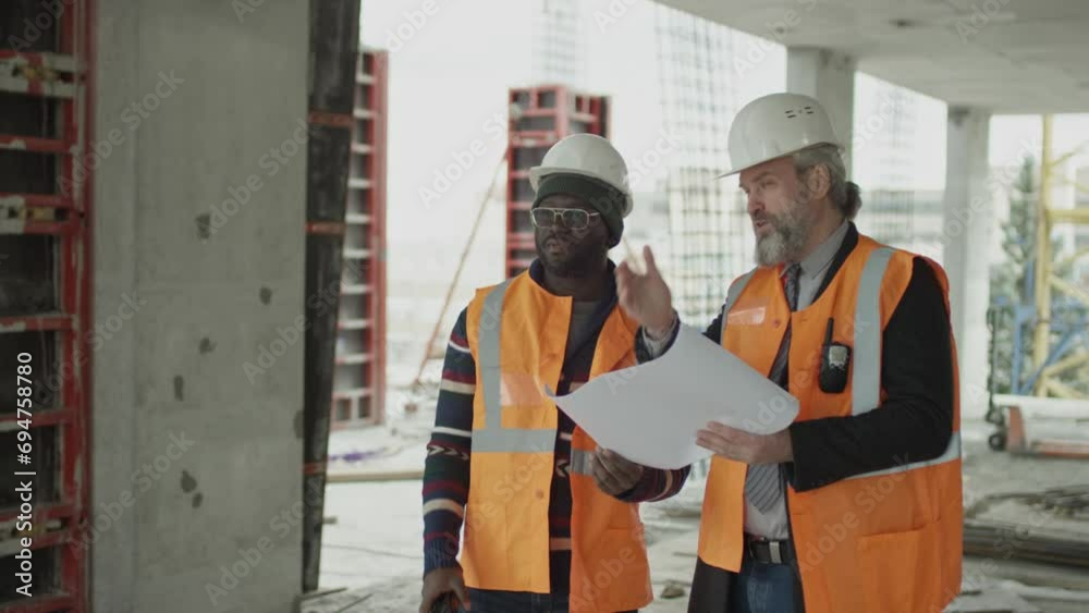 Medium long shot of two ethnically diverse male architects wearing safety vests and hardhats discussing blueprint at construction site Medium long shot of two ethnically diverse male architects wearing safety vests and hardhats discussing blueprint at construction site
