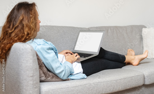 Rear view of woman sitting on sofa and working on laptop. Blank screen of laptop.