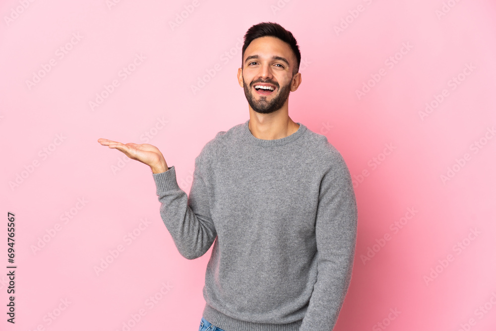 Young caucasian man isolated on pink background holding copyspace imaginary on the palm to insert an ad