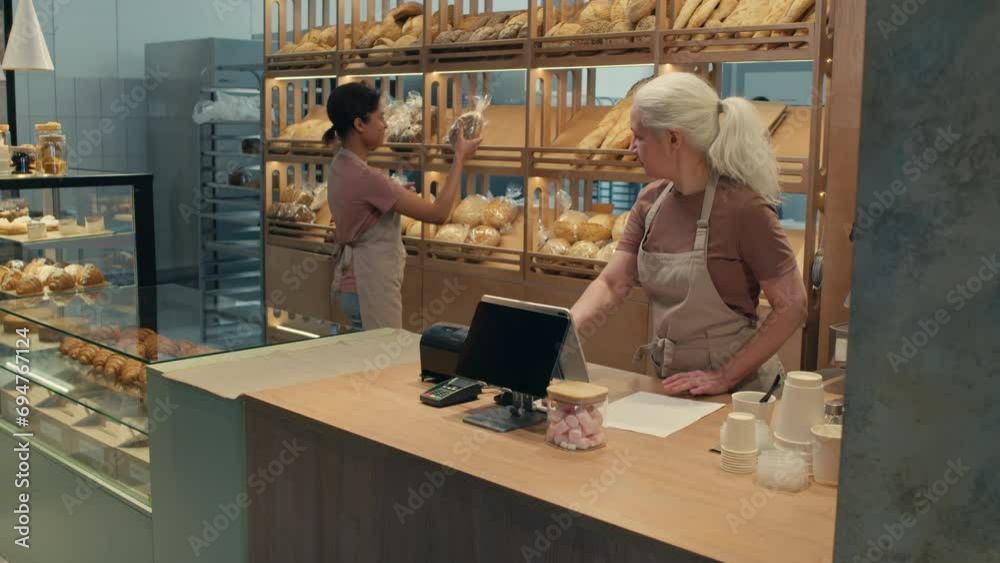 Young woman putting fresh bread on shelf, then going to cash register and helping senior colleague at work in bakery