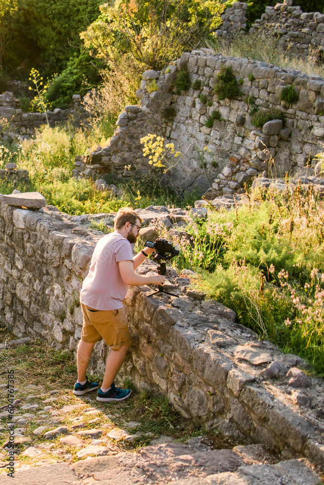 Cameraman in summer outfit with gimbal and cinema camera in his hands making travel video with old Europe Balkan nature and town on the background