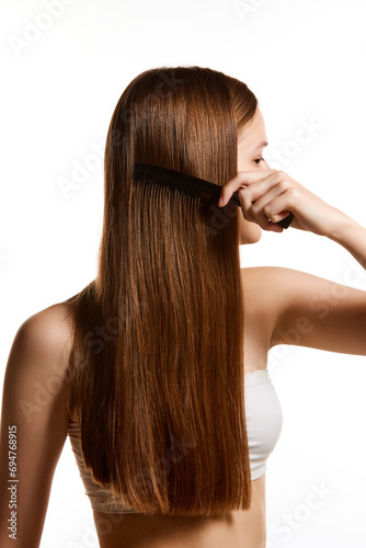 Young woman, female model with long silky straight brunette hair combing her hair against white studio background.