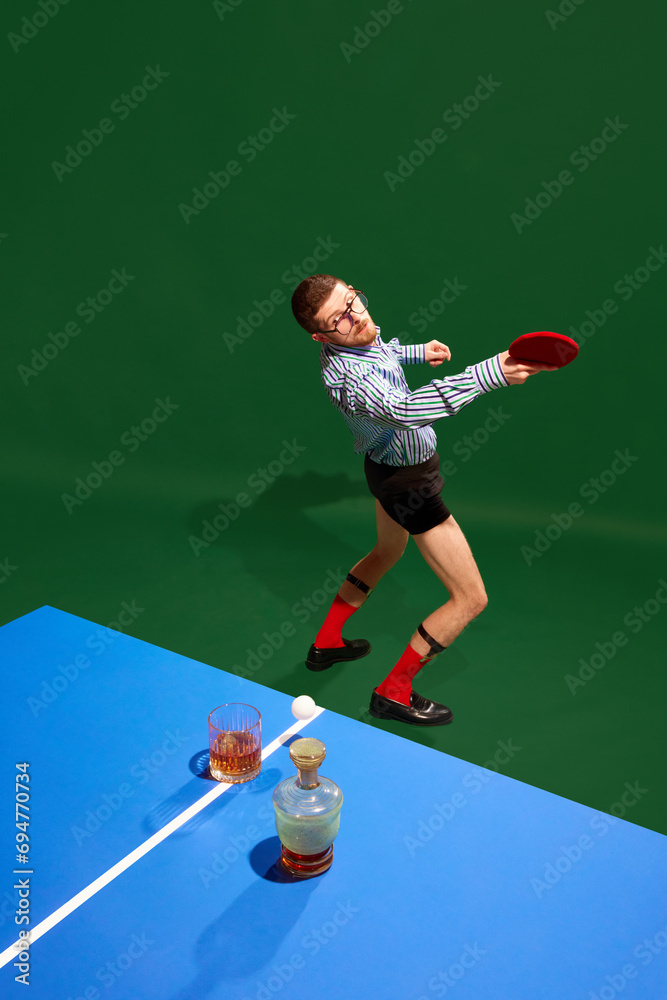 Top view. Concentrated young man in shirt and underwear playing table tennis with whiskey glass over green background. Concept of sport, leisure, hobby, creativity, fun and joy. Pop art Top view. Concentrated young man in shirt and underwear playing table tennis with whiskey glass over green background. Concept of sport, leisure, hobby, creativity, fun and joy. Pop art