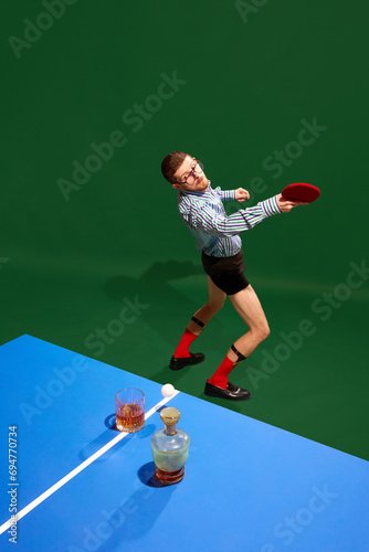 Top view. Concentrated young man in shirt and underwear playing table tennis with whiskey glass over green background. Concept of sport, leisure, hobby, creativity, fun and joy. Pop art © master1305