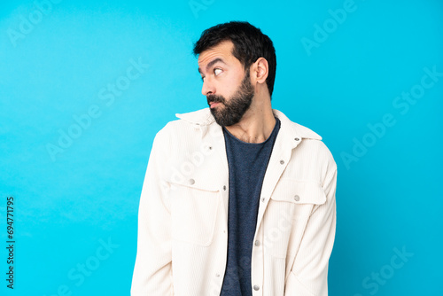 Young handsome man with white corduroy jacket over isolated blue background making doubts gesture looking side