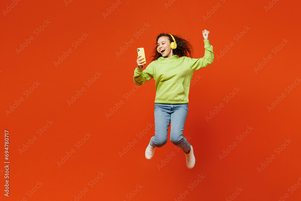 Full body young woman of African American ethnicity wear hoody casual clothes jump high hold use mobile cell phone listen music in headphones do winner gesture isolated on plain red orange background. Full body young woman of African American ethnicity wear hoody casual clothes jump high hold use mobile cell phone listen music in headphones do winner gesture isolated on plain red orange background.