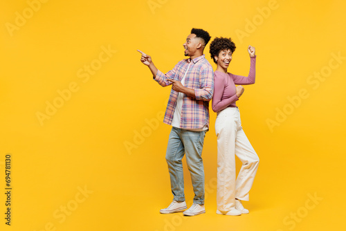Full body young happy couple two friends family man woman of African American ethnicity wearing casual clothes together point index finger aside do winner gesture isolated on plain yellow background.