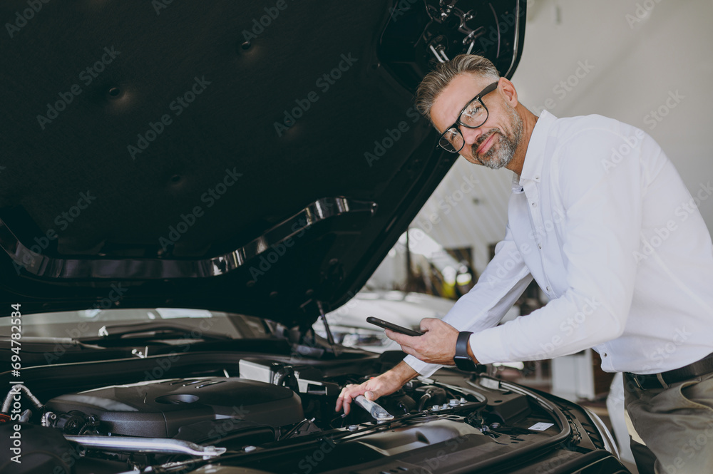 Adult man customer male buyer client he wear shirt examining engine open car hood use mobile cell phone choose auto want to buy new automobile in showroom vehicle salon store motor show Sales concept Adult man customer male buyer client he wear shirt examining engine open car hood use mobile cell phone choose auto want to buy new automobile in showroom vehicle salon store motor show Sales concept