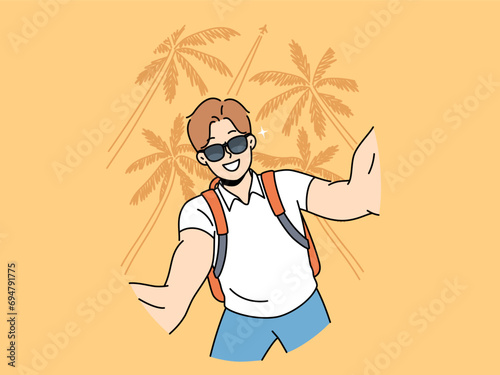Man tourist in sunglasses takes selfie traveling to hot resort or sunny beach with palm trees. Young guy tourist with backpack smiles photographing himself for blog about vacation trip