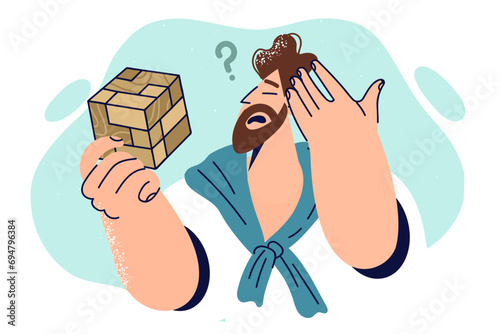Thoughtful man with wooden puzzle scratches head trying to solve difficult problem