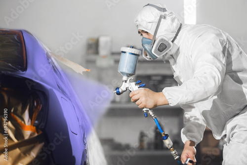 Male worker in protective clothes and mask painting car body to blue violet color using spray paint gun. 
