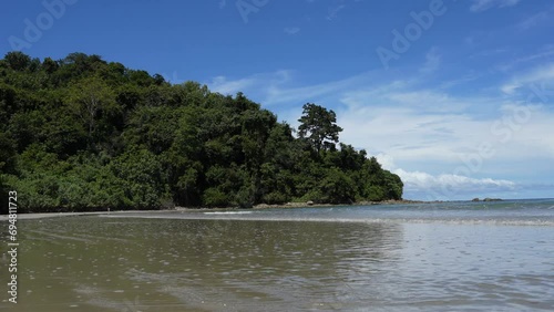 Summer on a tropical island. The waves of the turquoise ocean roll towards the shore, foaming and spreading along the beach. A hill  with green vegetation, against a blue sky, clouds. Reflection 