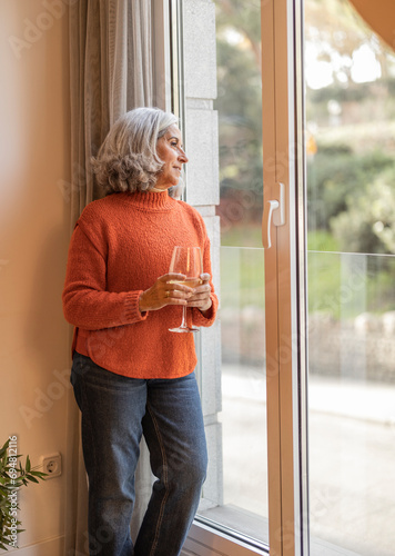 Lonely grandmother looking out the window while having a glass of wine waiting for the arrival of her family