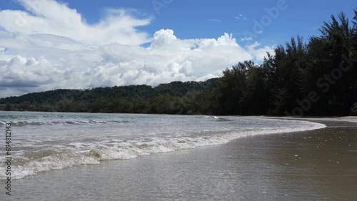 Surf in the ocean. Turquoise waves roll towards the shore, foam and spread over the sandy beach. Lush tropical vegetation on the shore. Clouds in the blue sky. Malaysia. Borneo. Kota Kinabalu 